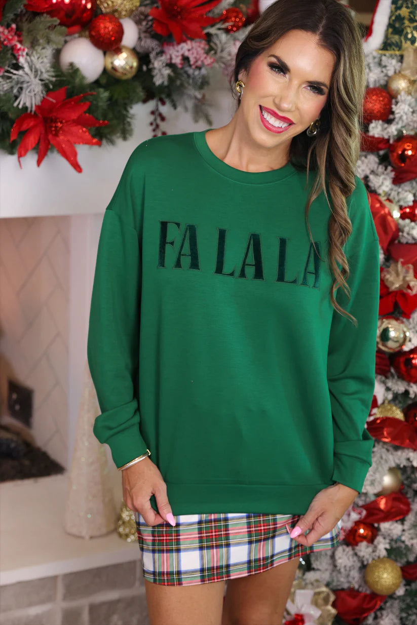 Woman wearing a green 'FA LA LA' sweater in front of a Christmas tree and fireplace.
