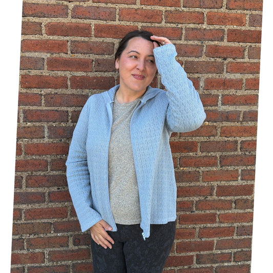 Woman wearing a light blue cardigan over a gray shirt, standing against a brick wall.